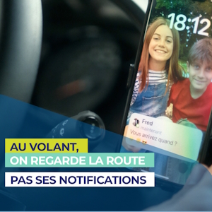 Description de l’image : On voit un smartphone fixé sur le tableau de bord d’une voiture. L’écran du téléphone affiche l’écran de verrouillage avec une photo de fond représentant une jeune fille souriante aux cheveux longs, portant une salopette en jean, aux côtés d’un garçon souriant en t-shirt rouge. L’heure indiquée est 18:12.  Une notification de message apparaît à l’écran. Le message provient d’un contact nommé Fred, qui écrit : « Vous arrivez quand ? ».  En bas à gauche de l’image, un message de sensibilisation est superposé dans trois encadrés de couleur (jaune, vert et bleu). Il est écrit : AU VOLANT, (en jaune)  ON REGARDE LA ROUTE (en vert)  PAS SES NOTIFICATIONS (en bleu foncé)  Sens du message : Il s’agit d’une campagne de prévention routière incitant les conducteurs à ne pas regarder leur téléphone lorsqu’ils conduisent. Le slogan rappelle que la priorité est la route, pas les notifications.